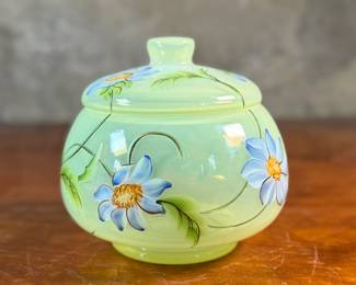 This lot features a Fenton "Dahlias" on Topaz Opalescent covered box, hand-painted by artist Frances Burton. Edition 386/1950 and measures, 5.5” H x 7” W x 7” D. The piece is crafted from Topaz Opalescent glass, which is a yellow mist color created by striking the opalescent glass before forming. The dahlia design features blue flowers with 22k gold highlights. The glass is also known as Vaseline glass and glows under a blacklight due to the use of depleted uranium in the glassmaking process.

