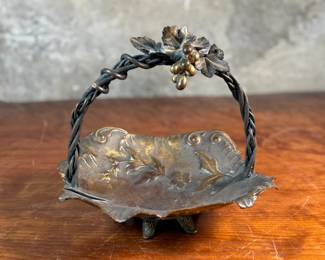 This lot features an American Aesthetic Movement trompe l’oeil basket, naturalistically modeled to simulate a cloth dish with a handle made of a bent twig and corded twine. The basket features gilt and silvered leaves and flower heads as decoration. The piece dates back to the 1940s–1950s and is made of solid brass. This piece measures, 7.5” H x 7.75” W x 7.75” D.

