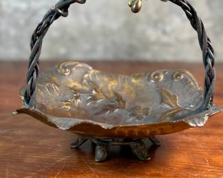 This lot features an American Aesthetic Movement trompe l’oeil basket, naturalistically modeled to simulate a cloth dish with a handle made of a bent twig and corded twine. The basket features gilt and silvered leaves and flower heads as decoration. The piece dates back to the 1940s–1950s and is made of solid brass. This piece measures, 7.5” H x 7.75” W x 7.75” D.

