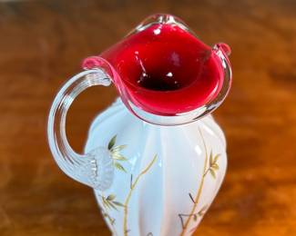 This lot features a Fenton Art Glass "Asian Garden" pitcher from the 2000 Connoisseur Collection. Limited edition 894/1850 and measures, 7.5” H x 5” W x 6” D. It features a hand-painted "Asian Garden" design on milk glass with a peach crest and a clear applied handle. The design was created by artist Robin Spindler and includes 22k gold highlights. 

