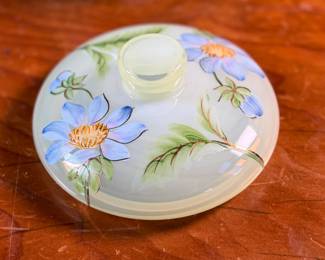 This lot features a Fenton "Dahlias" on Topaz Opalescent covered box, hand-painted by artist Frances Burton. Edition 386/1950 and measures, 5.5” H x 7” W x 7” D. The piece is crafted from Topaz Opalescent glass, which is a yellow mist color created by striking the opalescent glass before forming. The dahlia design features blue flowers with 22k gold highlights. The glass is also known as Vaseline glass and glows under a blacklight due to the use of depleted uranium in the glassmaking process.


