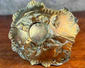 This lot features an American Aesthetic Movement trompe l’oeil basket, naturalistically modeled to simulate a cloth dish with a handle made of a bent twig and corded twine. The basket features gilt and silvered leaves and flower heads as decoration. The piece dates back to the 1940s–1950s and is made of solid brass. This piece measures, 7.5” H x 7.75” W x 7.75” D.

