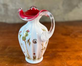 This lot features a Fenton Art Glass "Asian Garden" pitcher from the 2000 Connoisseur Collection. Limited edition 894/1850 and measures, 7.5” H x 5” W x 6” D. It features a hand-painted "Asian Garden" design on milk glass with a peach crest and a clear applied handle. The design was created by artist Robin Spindler and includes 22k gold highlights. 

