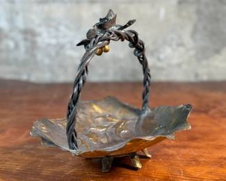 This lot features an American Aesthetic Movement trompe l’oeil basket, naturalistically modeled to simulate a cloth dish with a handle made of a bent twig and corded twine. The basket features gilt and silvered leaves and flower heads as decoration. The piece dates back to the 1940s–1950s and is made of solid brass. This piece measures, 7.5” H x 7.75” W x 7.75” D.

