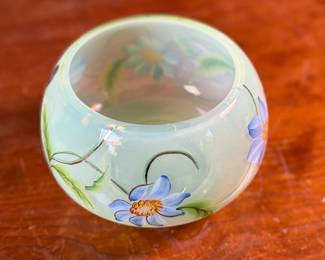 This lot features a Fenton "Dahlias" on Topaz Opalescent covered box, hand-painted by artist Frances Burton. Edition 386/1950 and measures, 5.5” H x 7” W x 7” D. The piece is crafted from Topaz Opalescent glass, which is a yellow mist color created by striking the opalescent glass before forming. The dahlia design features blue flowers with 22k gold highlights. The glass is also known as Vaseline glass and glows under a blacklight due to the use of depleted uranium in the glassmaking process.

