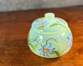 This lot features a Fenton "Dahlias" on Topaz Opalescent covered box, hand-painted by artist Frances Burton. Edition 386/1950 and measures, 5.5” H x 7” W x 7” D. The piece is crafted from Topaz Opalescent glass, which is a yellow mist color created by striking the opalescent glass before forming. The dahlia design features blue flowers with 22k gold highlights. The glass is also known as Vaseline glass and glows under a blacklight due to the use of depleted uranium in the glassmaking process.

