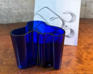 This lot features a Iittala Alvar Aalto Collection vase in ultramarine blue mouth-blown glass. It is also known as the Savoy Vase. The vase was designed by Finnish architect Alvar Aalto and his wife Aino for a design competition in 1936. It became an iconic piece of Finnish design after being displayed at the 1937 Paris World’s Fair. The unique, wavy shape was reportedly inspired by the hem of a Sami woman’s dress, with original sketches titled “The Eskimo Woman’s Leather Breeches”. Each vase is still manufactured by skilled glassblowers at the Iittala factory in Finland using original wooden molds. This vase is accompanied with Aalto’s MOMA book.

