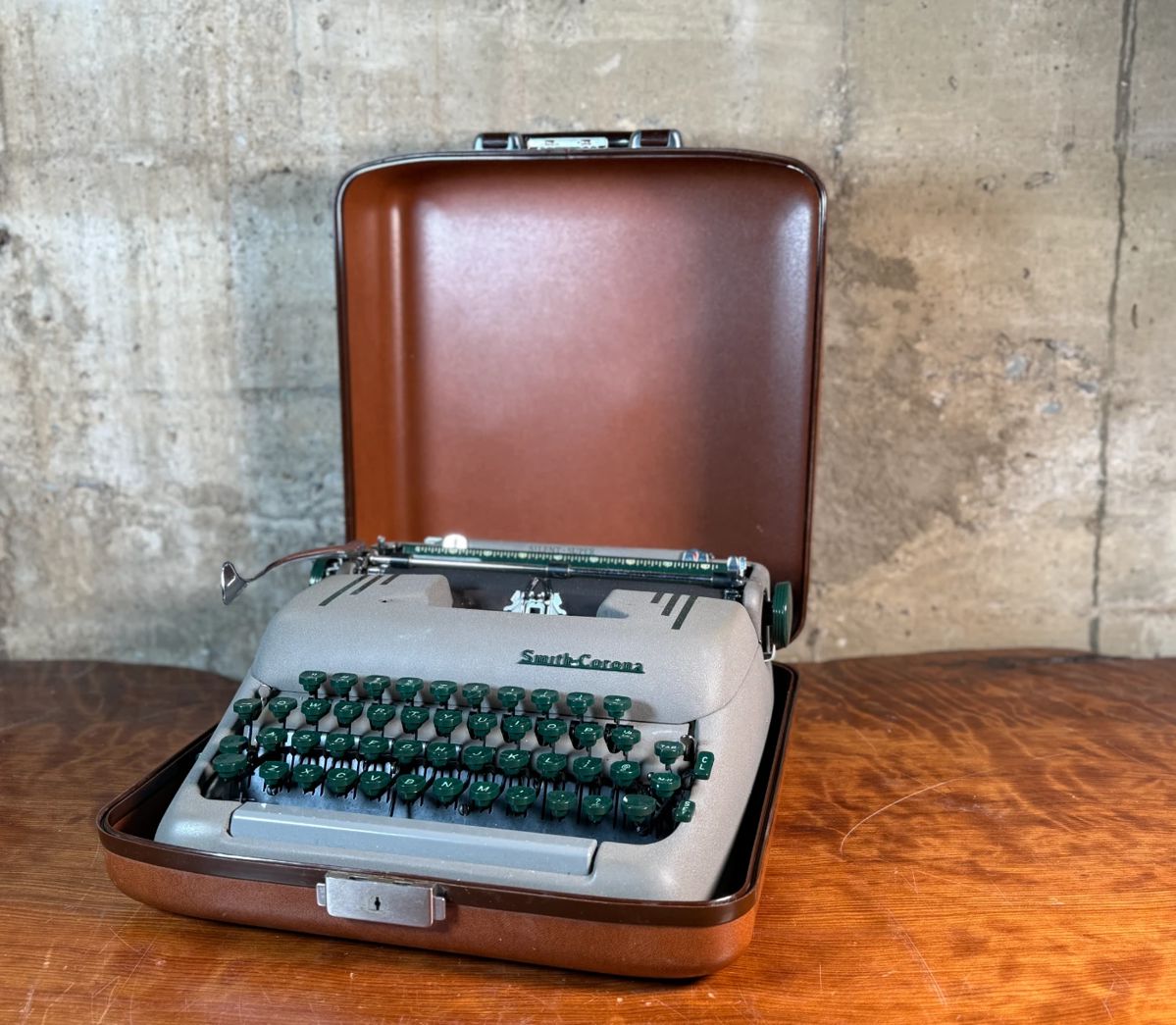 This lot features a vintage Smith Corona Silent-Super portable typewriter, circa 1950s. It has a khaki-toned body and forest green keys, and is shown with its original carrying case. 

