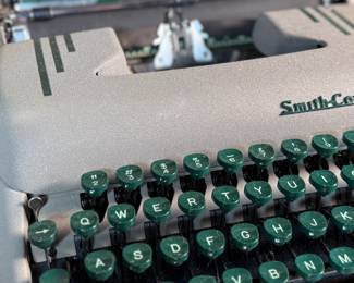 This lot features a vintage Smith Corona Silent-Super portable typewriter, circa 1950s. It has a khaki-toned body and forest green keys, and is shown with its original carrying case. 

