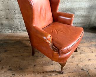 This lot features a Georgian-style leather wingback armchair in a warm, caramel or reddish-brown cowhide leather that measures, 42” H x 32” W x 27” D.

