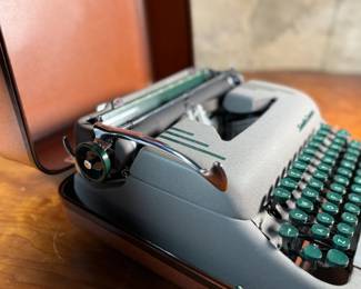 This lot features a vintage Smith Corona Silent-Super portable typewriter, circa 1950s. It has a khaki-toned body and forest green keys, and is shown with its original carrying case. 

