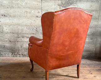 This lot features a Georgian-style leather wingback armchair in a warm, caramel or reddish-brown cowhide leather that measures, 42” H x 32” W x 27” D.

