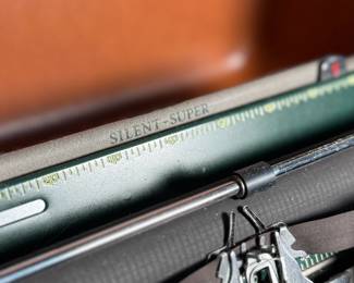 This lot features a vintage Smith Corona Silent-Super portable typewriter, circa 1950s. It has a khaki-toned body and forest green keys, and is shown with its original carrying case. 

