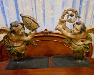 This is a pair of 18th-century Italian carved wooden cherubs.
They are hand-carved from wood and feature a parcel- painted finish with gold accents.
The cherubs present as reflections of each other with one holding a shield and the other a horn-like object.
