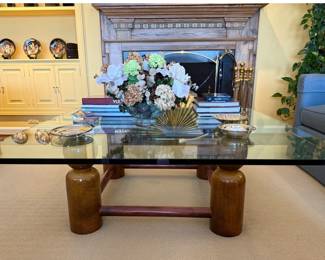 Large Glass-top Coffee Table:  Mid-Century Modern or Postmodern design featuring a thick glass top resting on a sculptural, solid wood base. The distinctive round, thick legs connected by a stretcher suggest a design that emphasizes the natural beauty of the wood and clean lines. 