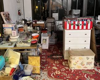 This photo shows a number of items: kitchen utensils, kitchen appliances, a red, white, and blue trunk, a white dresser, and too many others to list.