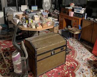 This photo shows another trunk, a desk, a classic-styled radio, a vacuum cleaner, and other electronic media devices.