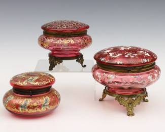 Three turn of the century Bohemian Glass dresser boxes.  Includes three circular Cranberry Glass boxes with Gilded metal mounts and floral enameled decoration.  Each unsigned, smallest with paper label from "The Burlington / N.Y.".  Medium box with area of damage at body and replaced hinge pin, some wear to decoration and Gilding.  Up to 5" high.   NOTE: From the estate of Bill & Barbara Shulhof of Saginaw, Michigan.
