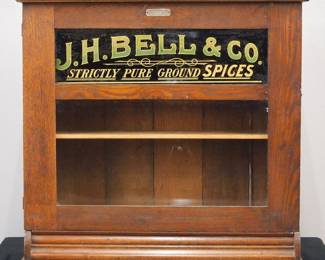 A turn of the century J. H. Bell store display cabinet.  Oak construction with hinged door and two glass panels, the upper with reverse painted advertising "J. H. Bell & Co, Strictly Pure Ground Spices", two interior shelves.  Original finish with some wear, some flaking to the reverse painted glass.  29 x 11 x 27 1/4" wide.  