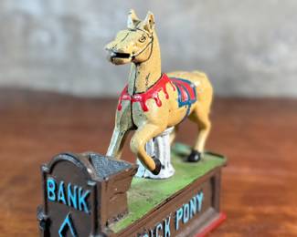 This lot features a reproduction cast iron mechanical bank known as the "Trick Pony". This bank measures, 7” H x 7” W x 2.5” D. The bank was originally patented in 1885 by Juleus Mueller, with manufacturing rights assigned to Charles and Walter Shepard. To operate the bank, a coin is placed in a small trough on the left, and a lever is pulled. 

