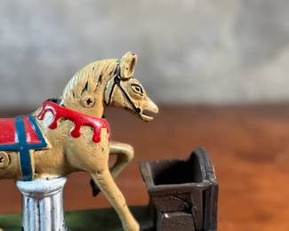 This lot features a reproduction cast iron mechanical bank known as the "Trick Pony". This bank measures, 7” H x 7” W x 2.5” D. The bank was originally patented in 1885 by Juleus Mueller, with manufacturing rights assigned to Charles and Walter Shepard. To operate the bank, a coin is placed in a small trough on the left, and a lever is pulled. 

