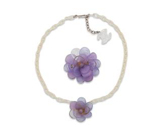 1158
CHANEL: A Set of Pâte de Verre Camélia Jewelry, 2001
A demi-parure featuring lavender purple glass camellias, comprising a choker necklace with a clear acrylic chain and CC charm to hook clasp, together with a large camellia pendant/brooch, each in silver toned metal, and each marked to plaque for Chanel, 2001 Printemps, Made in France

Necklace: 16 in.; Brooch: 2 in. diameter
44.5 grams gross
Estimate: $400 - $600