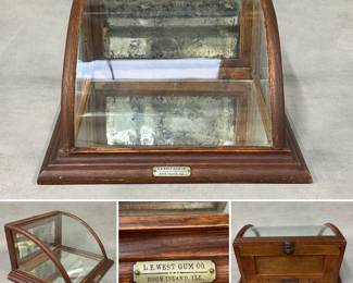 An antique curved glass countertop display case with a wooden frame, likely oak, with a curved glass front and mirrored back panels. Brass plate on front reads L.E. West Gum Co. Rock Island, Ill. 10" H x 17" W x 14" 