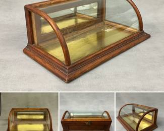 An antique curved glass countertop display case (one of three at this sale) with a wooden frame, likely oak, with a curved glass front and mirrored back panels. 7" H x 17.5" W x 10" D.