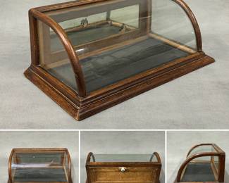 An antique curved glass countertop display case (one of three at this sale) with a wooden frame, likely oak, with a curved glass front and mirrored back panels. Painted gold lettering on top reads Jones & Hill Co.  7"H x 17-1/2" W x 10" D.