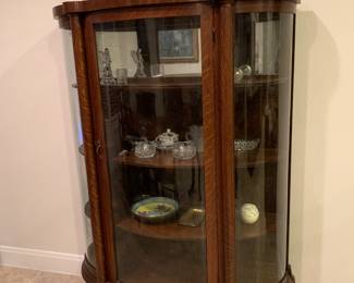 Antique Oak Curio Cabinet features rounded glass panels that wrap around its sides. It  offers a panoramic view of displayed items and it’s adorned with beautifully carved wood designs. 