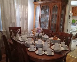 Wood dining room table with leaf and 6 wood chairs. Lighted wood China cabinet. Gibson white dinnerware set 62 pieces. Grace Concerto Fine China 47 pieces. Butter dishes. Glassware. 