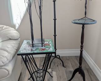Grape Motif Mosaic Side Table: A stunning stained glass and mosaic tile side table featuring a colorful grape vine design. It rests on a heavy wrought-iron base complete with a handy built-in magazine or book rack underneath.

Marble-Top Pedestal & Canopy Candle Holder: An elegant, tall wooden pedestal stand with a green marble-look top. Displayed perfectly on top is a beautiful wrought-iron candle holder featuring a unique canopy shade made of delicate metal leaves and dangling crystals.

Wrought Iron Floor Candelabras: A great collection of tall, heavy-duty black wrought-iron floor decor, including a sleek multi-tiered pillar candle stand and stunning branching tree sculptures equipped with glass votive cups.
