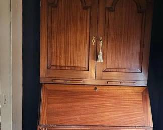 A Georgian style cherry wood two-part secretary, circa 19th century. The upper bookcase section with three turned finials on a broken arch bonnet above two paneled doors, that open to reveal a divided interior, with small drawers. The base portion with a slanted hinged lid that opens to a crafted interior, with an arrangement of drawers and cubbies, all over a serpentine front case having four drawers, mounted on a bracket base.