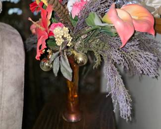 floral arrangement placed on a dark wooden accent table. The bouquet consists of a mix of faux tropical flowers, including what appear to be red hibiscus and pink calla lilies, interspersed with dry lavender fillers, artificial berries, and decorative feathers. The arrangement is housed in a slender, metallic-toned vase and is adorned with two small, gold-colored decorative spheres.
