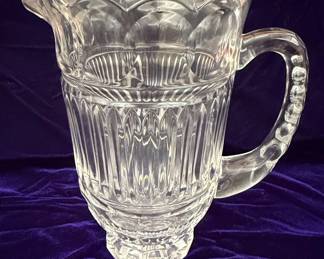A clear, pressed glass pitcher from Fifth Avenue Crystal, likely in their 'Princeton' pattern. The pitcher features a hobnail base, a body with vertical fluting and diamond-shaped panels, and a scalloped rim. The applied handle is decorated with raised dots. This pattern is characteristic of pressed glass from the late 19th or early 20th century.