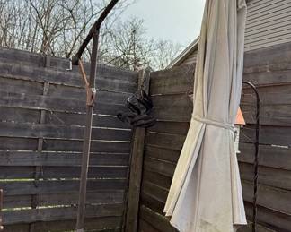 This is a cantilever or offset patio umbrella, characterized by its side-mounted pole that allows the canopy to extend over a seating or dining area without a central pole obstruction. The umbrella features a large, cream-colored fabric canopy, currently closed and tied. The supporting structure appears to be made of dark metal, likely aluminum or steel, with a rust-resistant coating. The base is a substantial, multi-piece cast iron construction designed to provide stability for the large umbrella. The individual base segments are decorative, featuring a fan-like or sunburst pattern. The overall style suggests a functional and decorative outdoor furnishing suitable for patios, decks, or gardens.