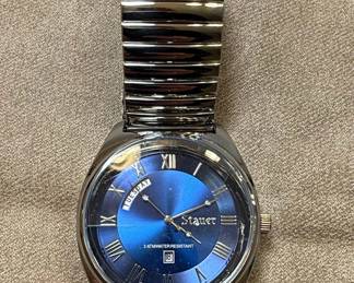 This Stauer men's watch features a striking blue sunburst dial with Roman numeral hour markers and a day-date complication. The case and expandable link bracelet are constructed from stainless steel, offering durability and a classic aesthetic. The watch is water-resistant to 3 ATM, suitable for everyday wear but not for swimming or diving. The dial is protected by a mineral crystal, and the case back displays the Stauer logo, a silhouetted craftsman at an anvil, along with 'STAINLESS STEEL BASE METAL' and '3 ATM WATER RESISTANT' markings. The movement is indicated as 'JAPAN MOVT' and a model number '13477' is present. The bracelet is identified as a 'Flex' style.