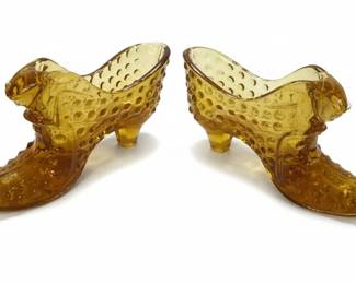 This pair of amber glass shoe slippers are a classic example of Fenton Art Glass, likely produced between the 1950s and 1970s. They are crafted from amber-colored glass, featuring a distinctive hobnail pattern across the body and a decorative cat's head on the vamp. The shoes are designed in a high-heeled, Victorian-esque style, complete with a pointed toe and a sculpted heel. The translucent amber hue allows light to pass through, highlighting the intricate hobnail texture and the molded details of the shoe's construction. These decorative pieces were popular collectibles during the mid-20th century, often used as decorative accents or small candy dishes. The presence of the Fenton oval logo on the sole confirms the maker.