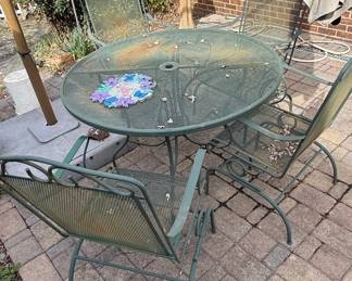 This vintage patio dining set features a round table and four chairs, all constructed from green-painted metal mesh. The table has a central umbrella hole and a decorative scrollwork base, while the chairs exhibit a classic design with curved backs and supportive frames. The mesh construction allows for water drainage and adds to the airy aesthetic. The set displays signs of age and use, including some surface rust and wear on the paint, consistent with outdoor exposure over time. It embodies a timeless, functional style often associated with mid-20th century outdoor furniture.
