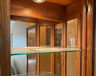 This is a traditional corner curio cabinet constructed from oak wood, featuring a multi-sided glass display area. The cabinet has a stepped top with decorative molding and a mirrored back panel, visible through the glass. It includes several adjustable glass shelves for displaying collectibles. A single interior light fixture is present at the top of the cabinet. The overall style suggests a late 20th-century or early 21st-century design, likely intended for residential use to showcase decorative items. The wood appears to be solid oak or oak veneer, with a medium-tone stain.