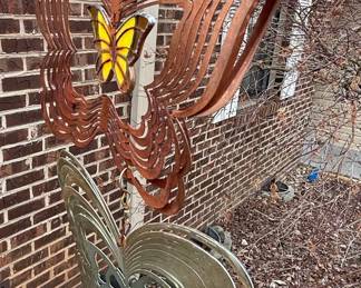 This kinetic metal wind spinner features a central floral-like design with layered, scalloped metal petals in a distressed yellow and gold finish. The center of the spinner is adorned with a circular panel depicting a butterfly and a blue sky with clouds, rendered in a painted or enameled style. Below the main spinner, a tiered arrangement of layered metal butterfly forms are suspended, with the top butterfly in a rusted copper patina and the bottom in a brushed silver finish. The overall aesthetic suggests a whimsical garden art piece designed to rotate and move with the wind, creating a dynamic visual display. The construction appears to be of durable metal, suitable for outdoor display, with elements of glass or enamel for the central butterfly detail. The item is characterized by its kinetic movement and the inclusion of multiple butterfly motifs, with varying metal finishes.