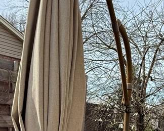 This is a cantilever or offset patio umbrella, characterized by its side-mounted pole that allows the canopy to extend over a seating or dining area without a central pole obstruction. The umbrella features a large, cream-colored fabric canopy, currently closed and tied. The supporting structure appears to be made of dark metal, likely aluminum or steel, with a rust-resistant coating. The base is a substantial, multi-piece cast iron construction designed to provide stability for the large umbrella. The individual base segments are decorative, featuring a fan-like or sunburst pattern. The overall style suggests a functional and decorative outdoor furnishing suitable for patios, decks, or gardens.