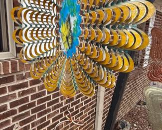 This kinetic metal wind spinner features a central floral-like design with layered, scalloped metal petals in a distressed yellow and gold finish. The center of the spinner is adorned with a circular panel depicting a butterfly and a blue sky with clouds, rendered in a painted or enameled style. Below the main spinner, a tiered arrangement of layered metal butterfly forms are suspended, with the top butterfly in a rusted copper patina and the bottom in a brushed silver finish. The overall aesthetic suggests a whimsical garden art piece designed to rotate and move with the wind, creating a dynamic visual display. The construction appears to be of durable metal, suitable for outdoor display, with elements of glass or enamel for the central butterfly detail. The item is characterized by its kinetic movement and the inclusion of multiple butterfly motifs, with varying metal finishes.