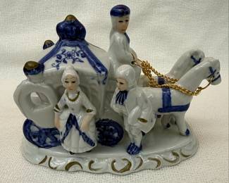 This is a delicate blue and white porcelain figurine depicting a horse-drawn carriage with three figures. The carriage is adorned with blue floral motifs, characteristic of traditional European porcelain designs. The figures, dressed in period attire, appear to be a driver and two passengers. The overall aesthetic suggests a Rococo or Neoclassical influence, common in decorative porcelain pieces from the 19th and 20th centuries. The gold-colored chain connecting the horses to the carriage adds a touch of opulence. The base is sculpted with decorative elements, enhancing its display value. The blue and white color scheme is a prominent feature.