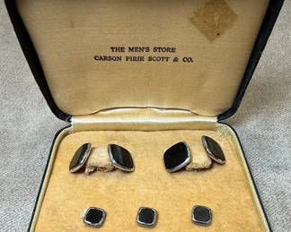 This is a vintage set of cufflinks and shirt studs presented in their original presentation box from "The Men's Store, Carson Pirie Scott & Co.". The set includes three shirt studs and four cufflinks, with the cufflinks featuring a distinctive rounded diamond shape and the studs being square. Both the cufflinks and studs appear to be crafted from black onyx set within a silver-tone metal bezel. The presentation box is covered in black faux leather with a gold trim and lined with a tan velvet-like material. The interior lid bears the embossed branding of the retailer, indicating a mid-20th century origin, a period when such sets were common formal wear accessories.