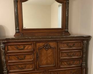 This bedroom set, comprising a tall chest of drawers, a nightstand, and a dresser with a mirror, is crafted from cherry wood with a rich, warm finish. The style is reminiscent of Italian Baroque, characterized by ornate carvings, scrolled hardware, and decorative inlays. Each piece features faux marble columns with detailed capitals and bases, adding a touch of opulence. The dresser top and nightstand top appear to be made of real marble, likely black with white veining, providing a durable and luxurious surface. The intricate detailing on the drawer fronts and the central decorative panel of the dresser suggests a high level of craftsmanship, typical of furniture designed to make a statement.