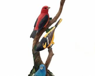 This Danbury Mint "Summer Serenade" figurine features four distinct bird species perched on a textured tree branch. The birds depicted are a Scarlet Tanager, an Oriole, a Bluebird, and a smaller yellow and gray bird, possibly a warbler or vireo. The base has mossy textures and floral accents. The figurine is made of resin and meticulously painted to showcase the birds' natural beauty and detail.