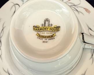 This set includes a lidded sugar bowl, a creamer pitcher, and a teacup with its matching saucer, all crafted from fine china. The pattern, "Platinum Garland," features delicate platinum trim along the edges and a graceful design of wheat stalks and leaves in a muted gray and subtle copper hue. The pieces are white with a smooth, glossy finish, indicative of quality porcelain. The sugar bowl has elegant, curved handles and a decorative lid finial. The creamer exhibits a classic spout and a single handle, while the teacup has a matching handle and sits on a saucer adorned with the same garland motif. The maker's mark on the underside of the teacup identifies it as Harmony House Fine China, Platinum Garland, Made in Japan, with the pattern number 3541.