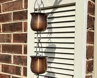 This is a decorative and functional rain chain made of copper, featuring a series of cup-shaped elements linked together by chains. The cups are designed to guide rainwater from the gutter to the ground, creating a visually appealing water flow. The copper material will develop a natural patina over time, adding to its aesthetic appeal. The overall style is classic and suitable for traditional or contemporary home exteriors. The cups appear to be of a simple, smooth design.