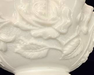 A vintage white milk glass bowl by Imperial Glass, featuring an embossed "American Beauty" rose pattern with leaves. The rim is scalloped and ruffled, with a crimped edge. The bowl has a satin finish and sits on a low pedestal base. This piece is characteristic of mid-century American glassware, often used as a rose bowl or decorative centerpiece.